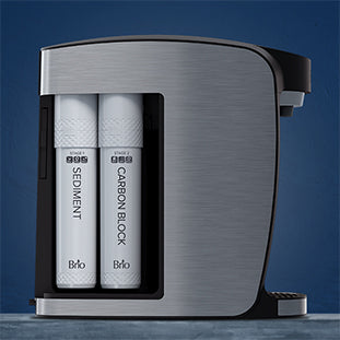 A sleek, silver countertop water filtration system with two visible filter cartridges labeled Sediment and Carbon Block against a blue background.